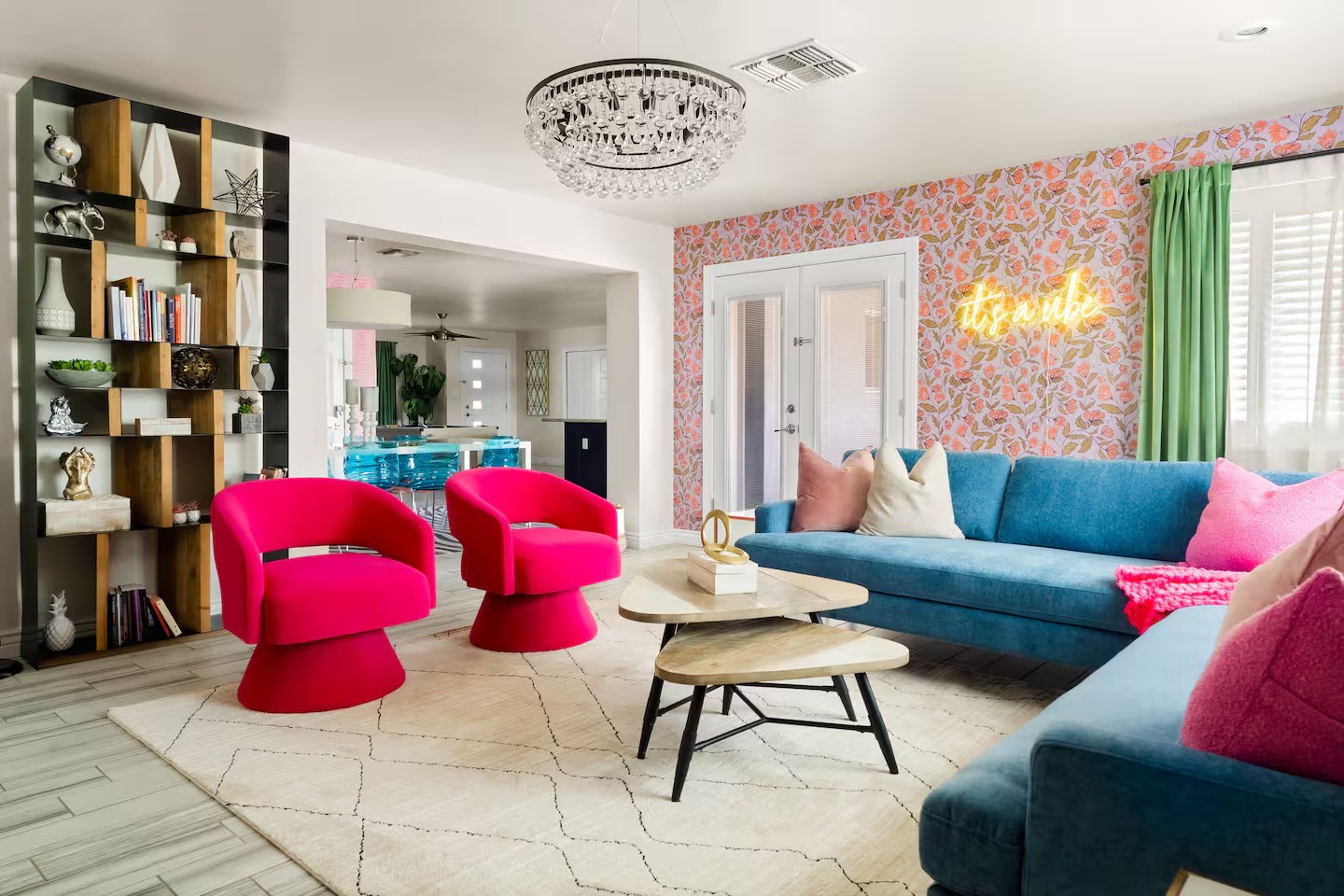 Living room with blue velvet sectional, two hot pink swivel chairs, crystal chandelier, coral floral wallpaper with yellow neon sign, and open timber shelving.