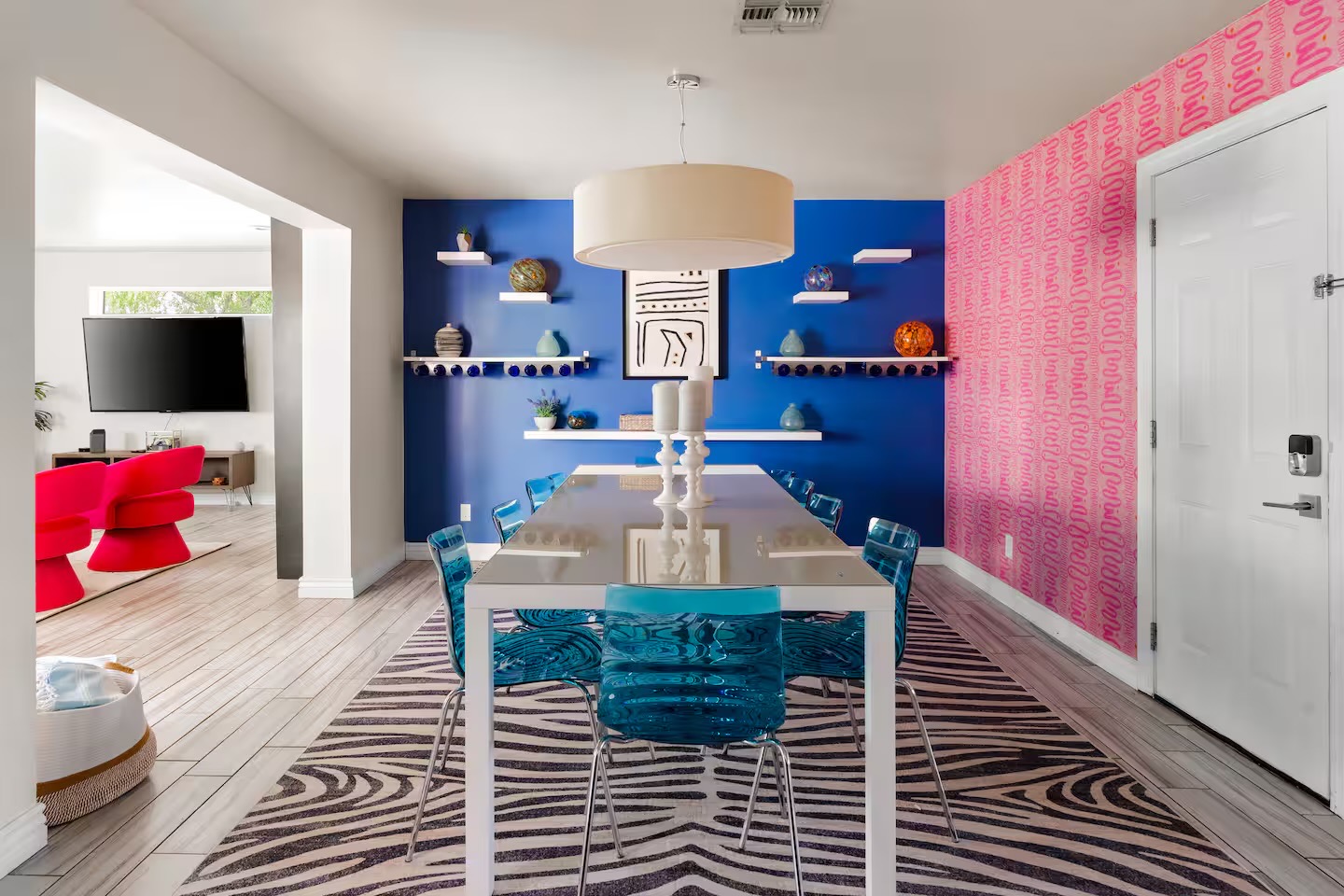 Dining room with white table, translucent teal chairs, zebra-print rug, drum pendant, cobalt accent wall with floating shelves, and pink patterned wallpaper.