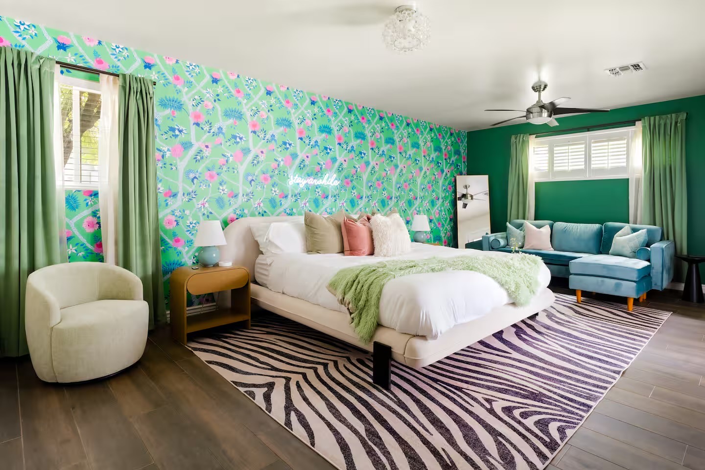 Bedroom with green chinoiserie floral wallpaper, emerald walls, cream upholstered bed, zebra-print rug, boucle swivel chair, and teal velvet sofa with ottoman.