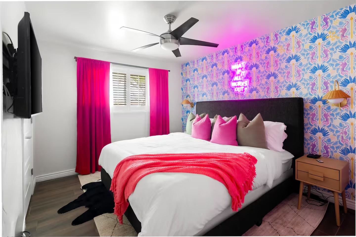 Bedroom with blue and lilac patterned feature wall, pink neon sign, dark upholstered bed, hot pink curtains and throw, and amber wall sconces.