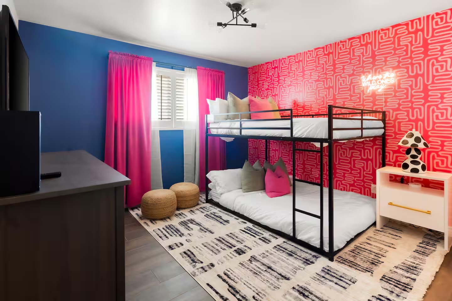 Bunk room with black metal bunk bed, coral maze-pattern wallpaper, cobalt blue walls, magenta curtains, jute poufs, and a black-and-cream striped rug.
