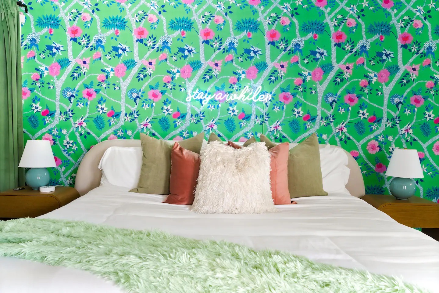 Bed detail with green and pink chinoiserie wallpaper, white neon script sign, layered sage and blush pillows, teal ceramic lamps, and a mint shag throw.