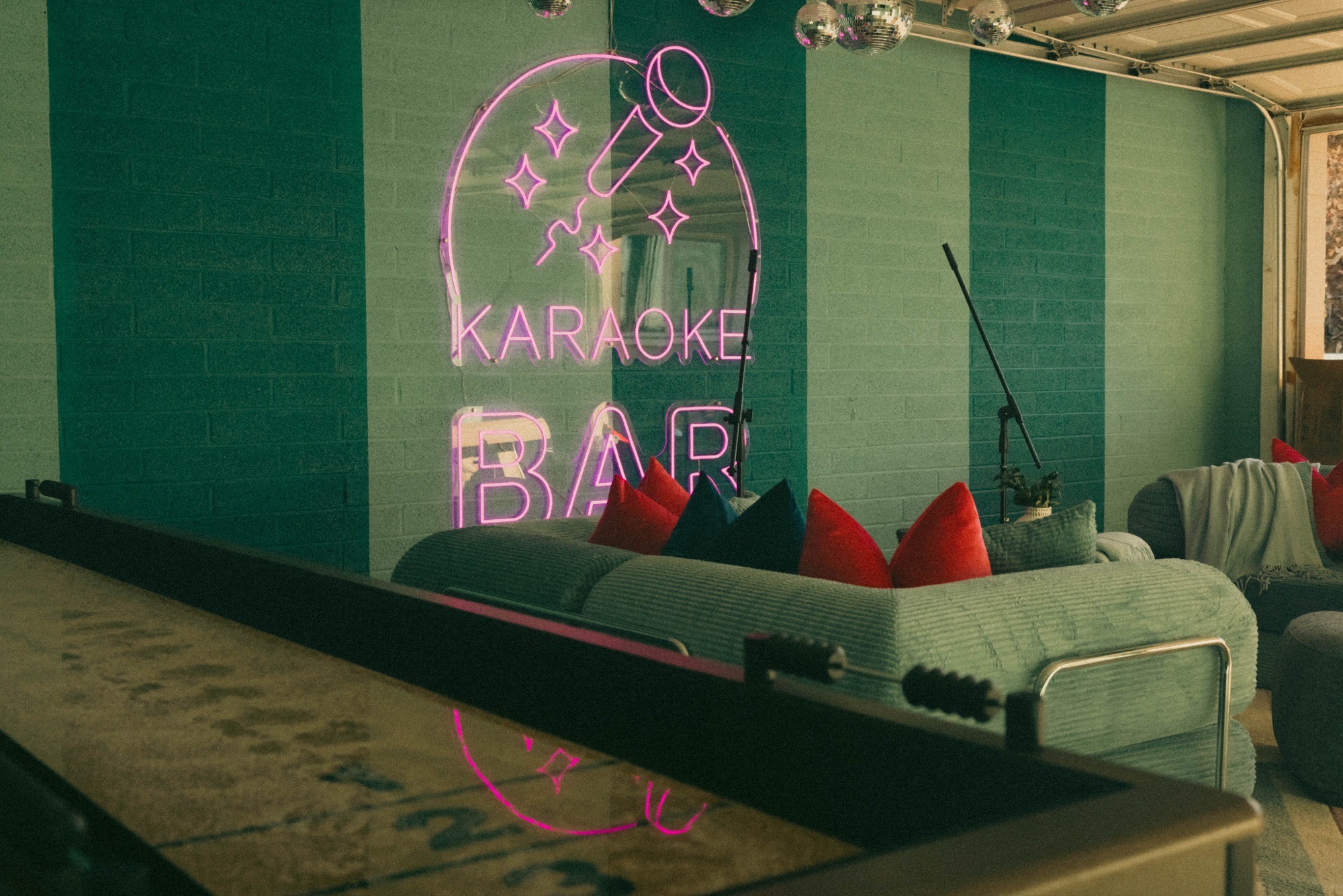 Karaoke lounge with teal-striped brick walls, pink neon sign, disco balls, sage corduroy sofa with red and teal pillows, shuffleboard table foreground.