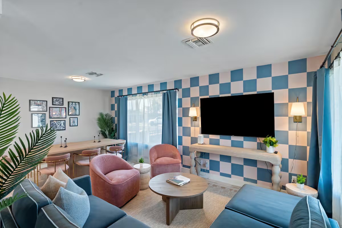 Living room with a blue and blush checkerboard accent wall, wall-mounted TV over a sculpted console, pink velvet swivel chairs and blue sofas.