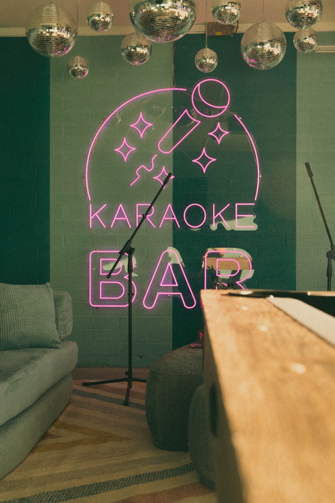 Karaoke lounge with pink neon Karaoke Bar sign on teal-striped brick, hanging disco balls, mic stands, sage sofa and wood bar top.