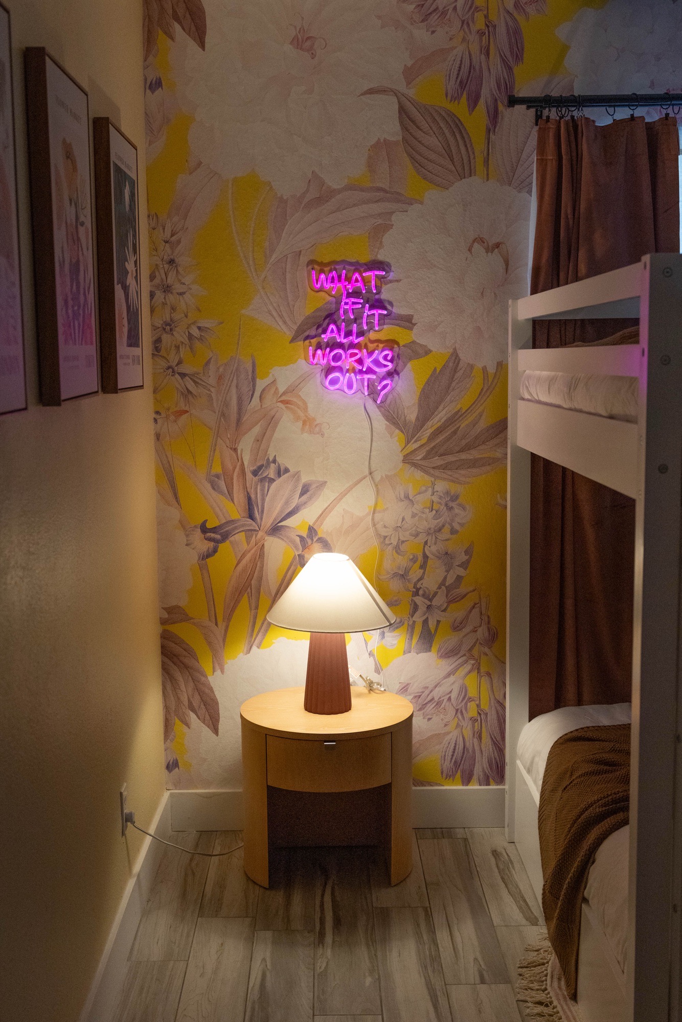 Bunk room nook with yellow oversized-floral wallpaper, pink neon script sign, ribbed terracotta lamp on round wood nightstand, white bunk bed, rust curtains.