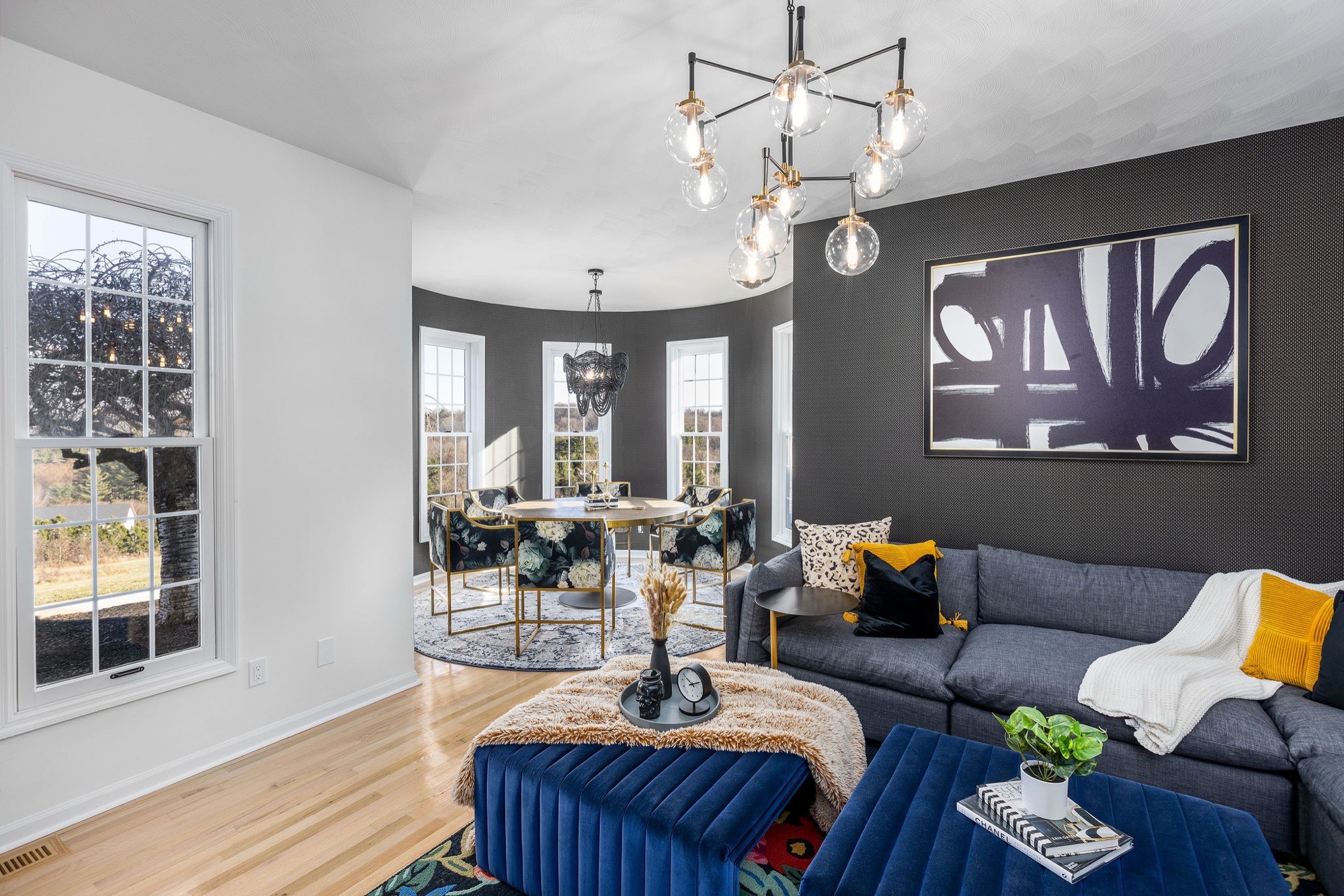 Living room with charcoal textured wallpaper, grey sectional, navy velvet ottomans and a globe chandelier, opening to a round dining turret.