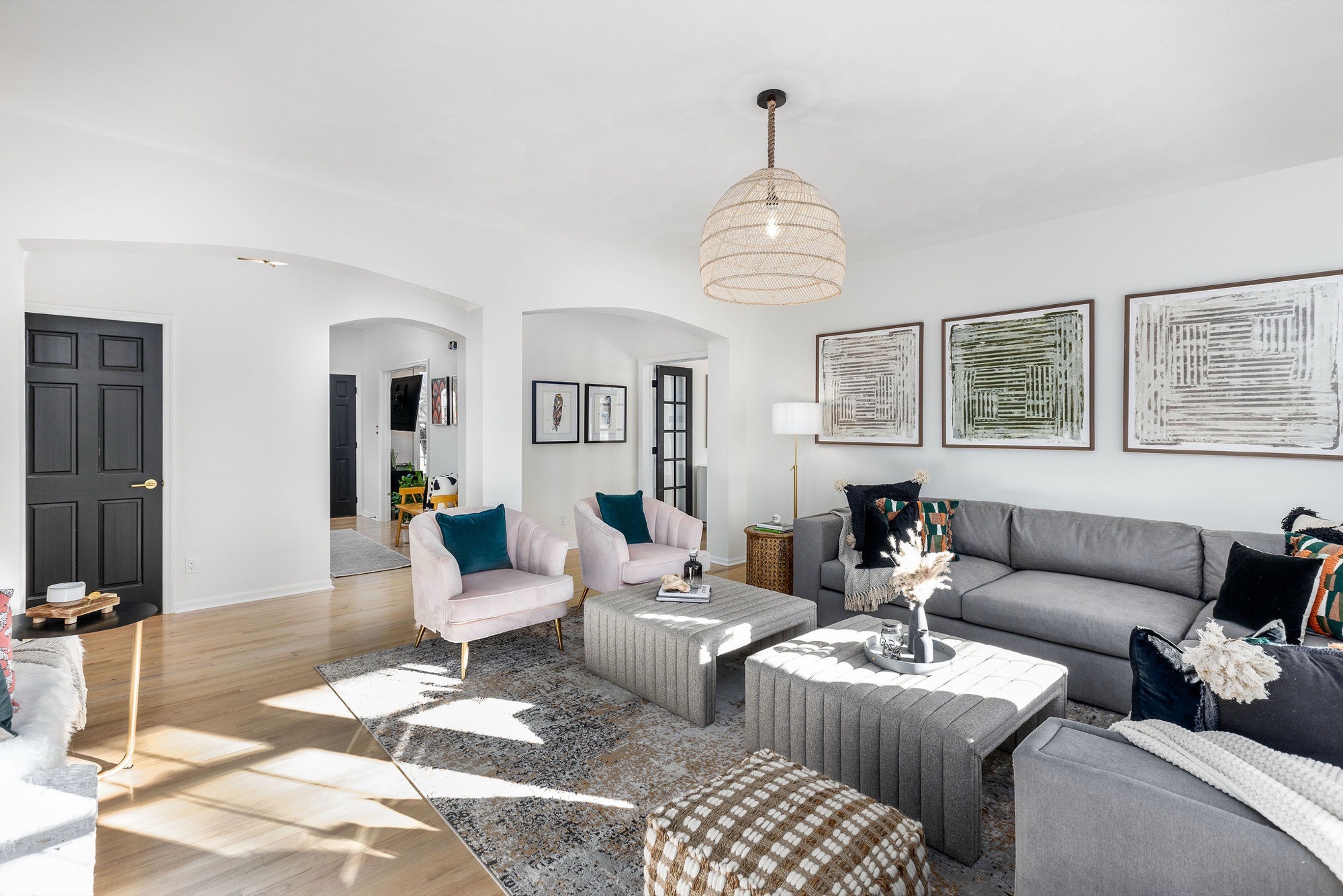 Bright living room with grey sofa, blush armchairs with teal pillows, grey upholstered ottomans, woven rattan pendant, abstract art and arched doorways.