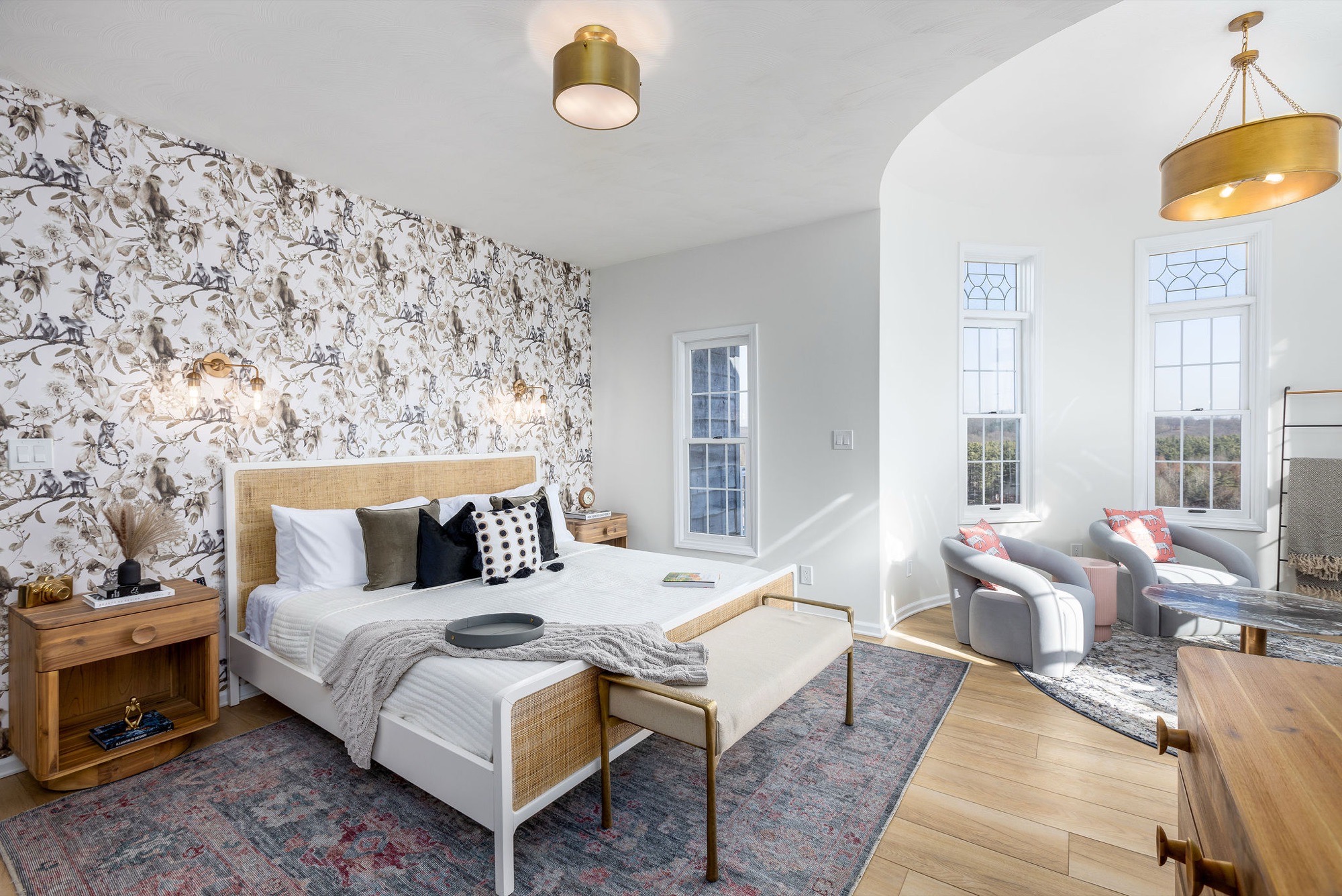 Bedroom with bird-and-floral wallpaper, white cane bed, brass sconces and drum pendant, plus a curved window nook with grey lounge chairs.