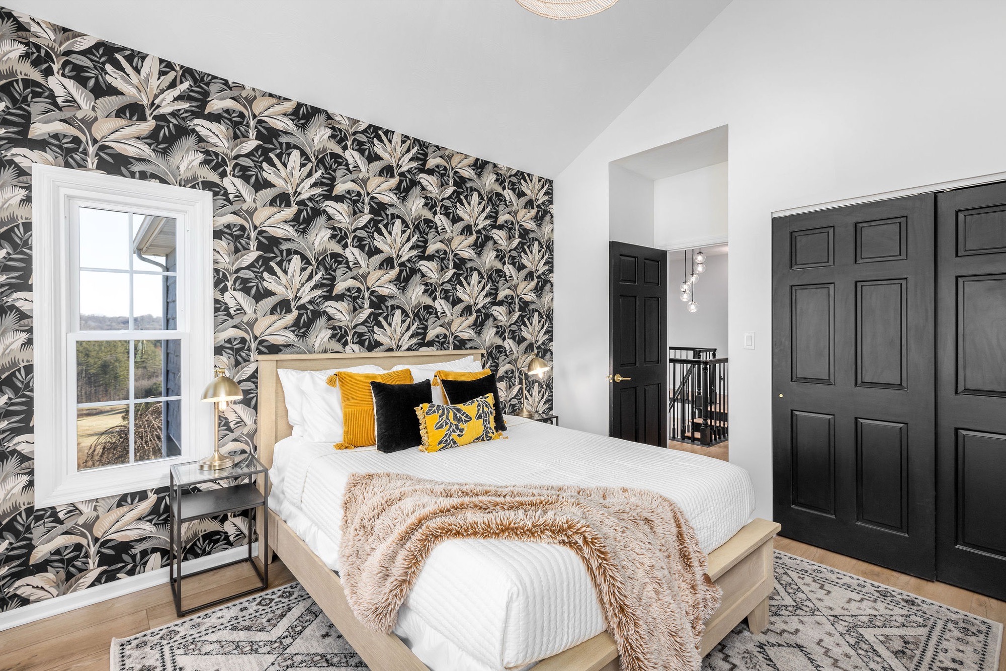 Vaulted bedroom with black-and-cream palm leaf wallpaper, pale wood bed, mustard and black pillows, brass lamp and black paneled doors.