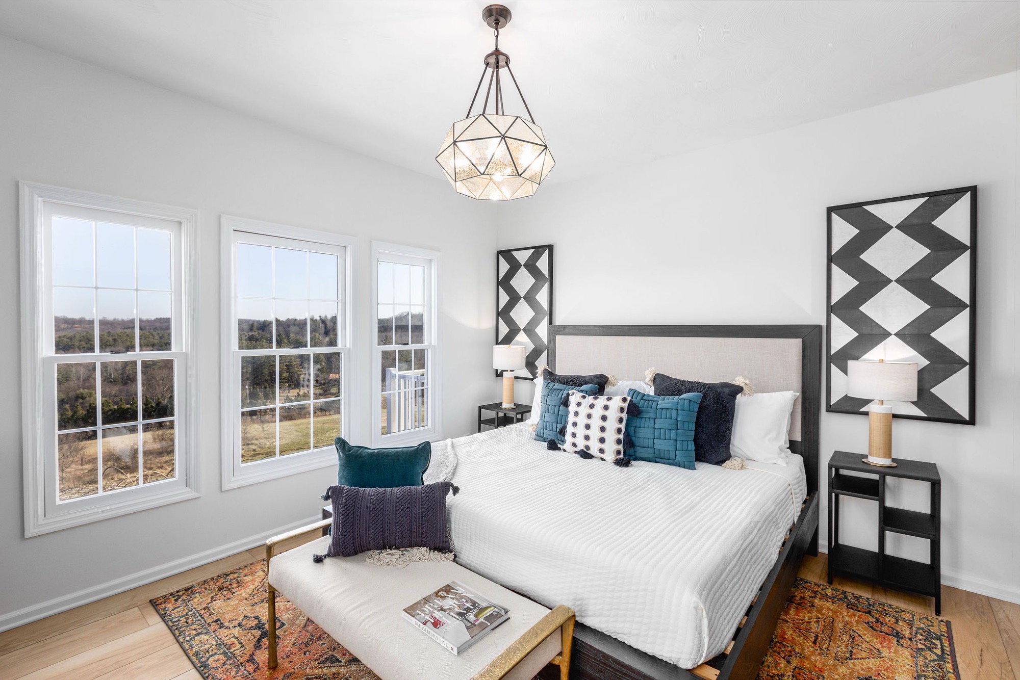 Bedroom with white walls, black-and-white geometric art panels, faceted glass pendant, upholstered bed with teal and navy pillows, and orange patterned rug.