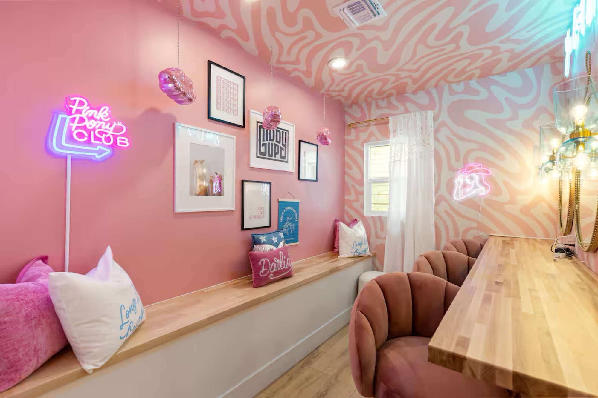 Pink lounge with swirl-patterned ceiling and wall, neon signs, gallery wall above a built-in timber bench, and blush velvet scalloped chairs at a butcher-block counter.
