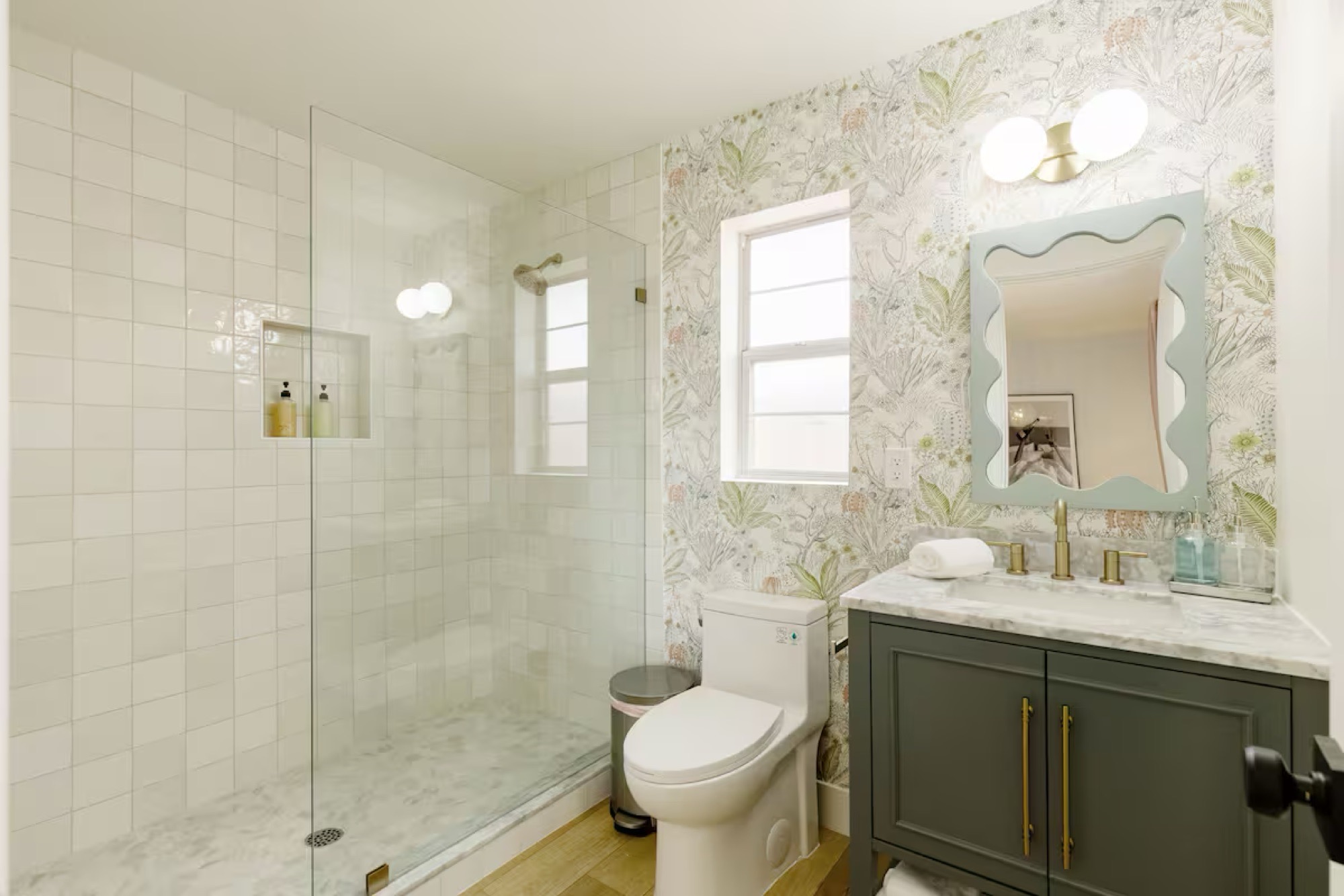 Bathroom with botanical wallpaper, wavy pale-blue mirror, dark green vanity with marble top and brass hardware, globe sconces and frameless glass shower in white tile.