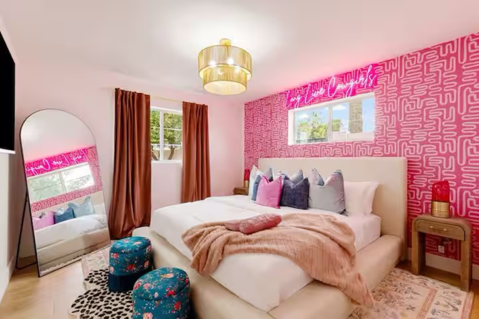 Bedroom with hot-pink maze-pattern accent wall, neon script sign, gold drum pendant, cream upholstered bed, rust velvet curtains, arched floor mirror and floral teal ottomans.