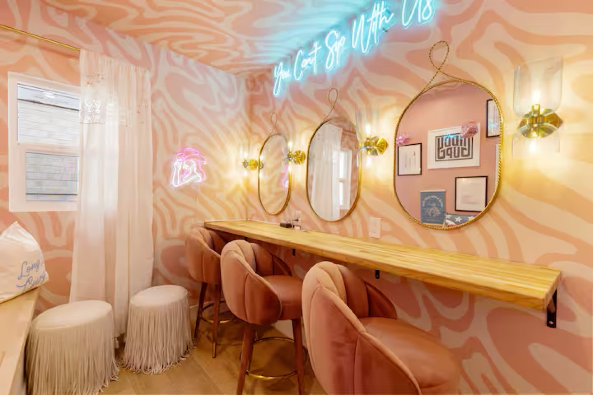 Vanity nook with pink-and-cream swirl walls, three gold-framed oval mirrors, brass sconces, long butcher-block counter, blush velvet stools and fringed white poufs beneath neon lettering.