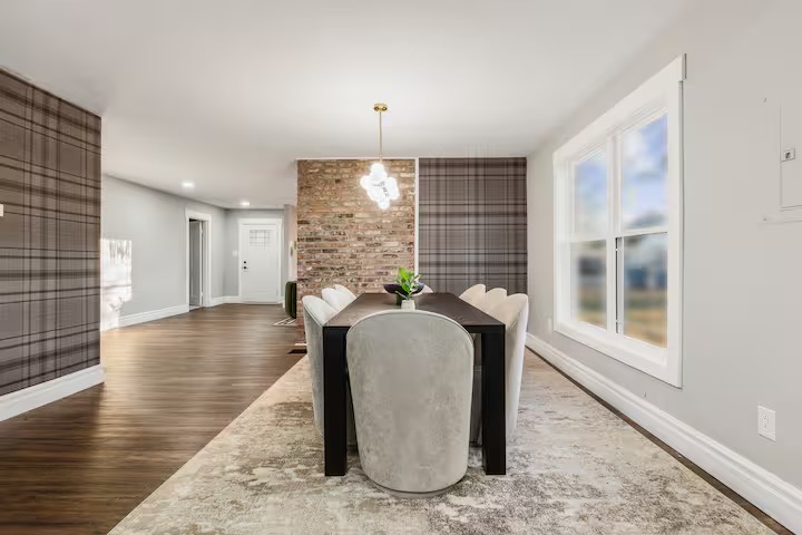 Dining area with a dark wood table, pale velvet scalloped-back chairs, exposed brick wall, taupe plaid wallpaper and a glass globe cluster pendant.
