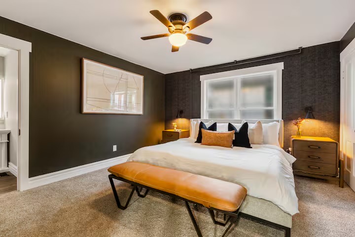 Bedroom with charcoal walls and textured wallpaper, white bedding, navy and cognac pillows, wood nightstands, black wall sconces and a cognac leather bench.
