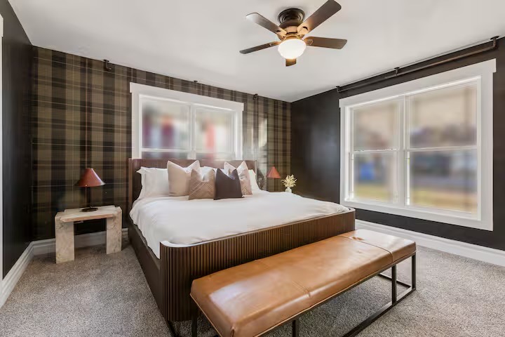 Bedroom with olive-brown plaid wallpaper, fluted wood bed frame, travertine side table, rust-shaded lamps and a cognac leather bench at the foot.
