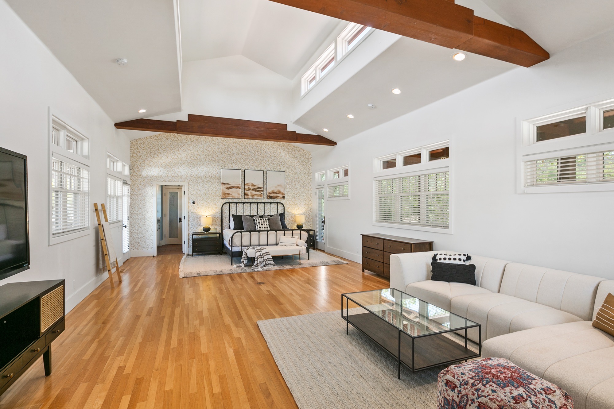 Expansive primary suite with vaulted white ceiling, dark timber beams, clerestory windows, honey wood floors, cream curved sectional and black iron bed.