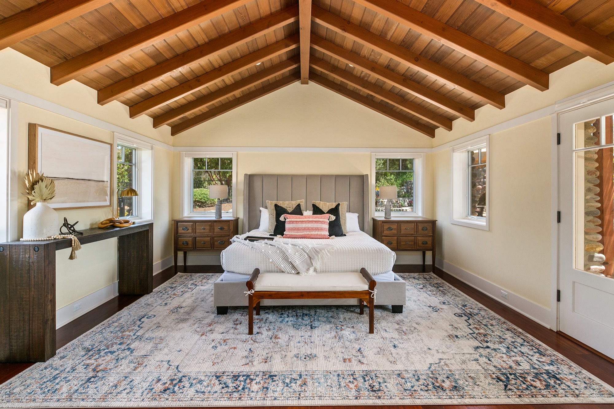 Bedroom under a vaulted tongue-and-groove wood ceiling with exposed beams, gray wingback bed, wooden nightstands and a blue-and-cream vintage-style rug.