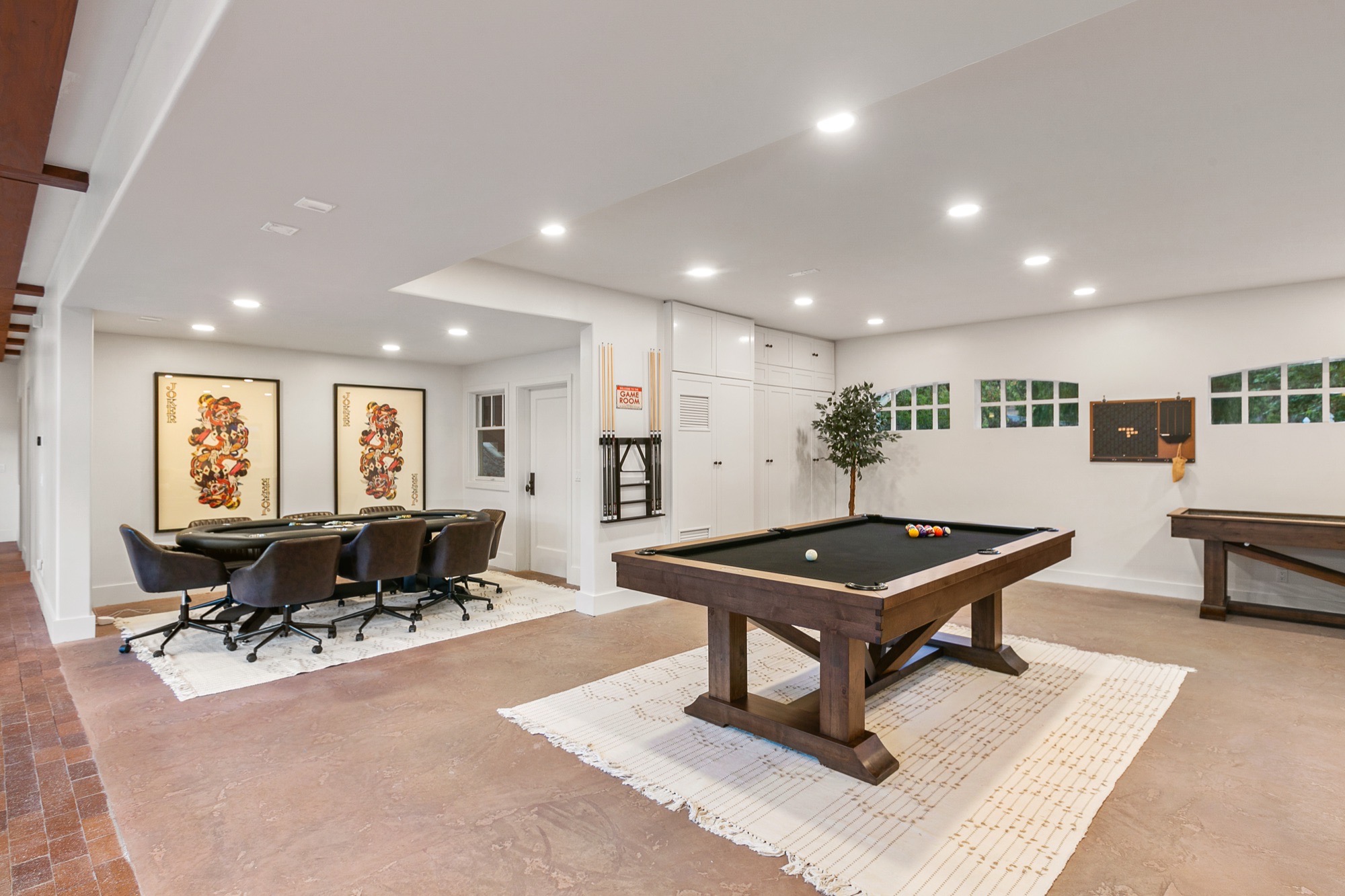 Game room with black-felt pool table on a wooden trestle base, oval card table with dark swivel chairs, framed art and recessed lighting.