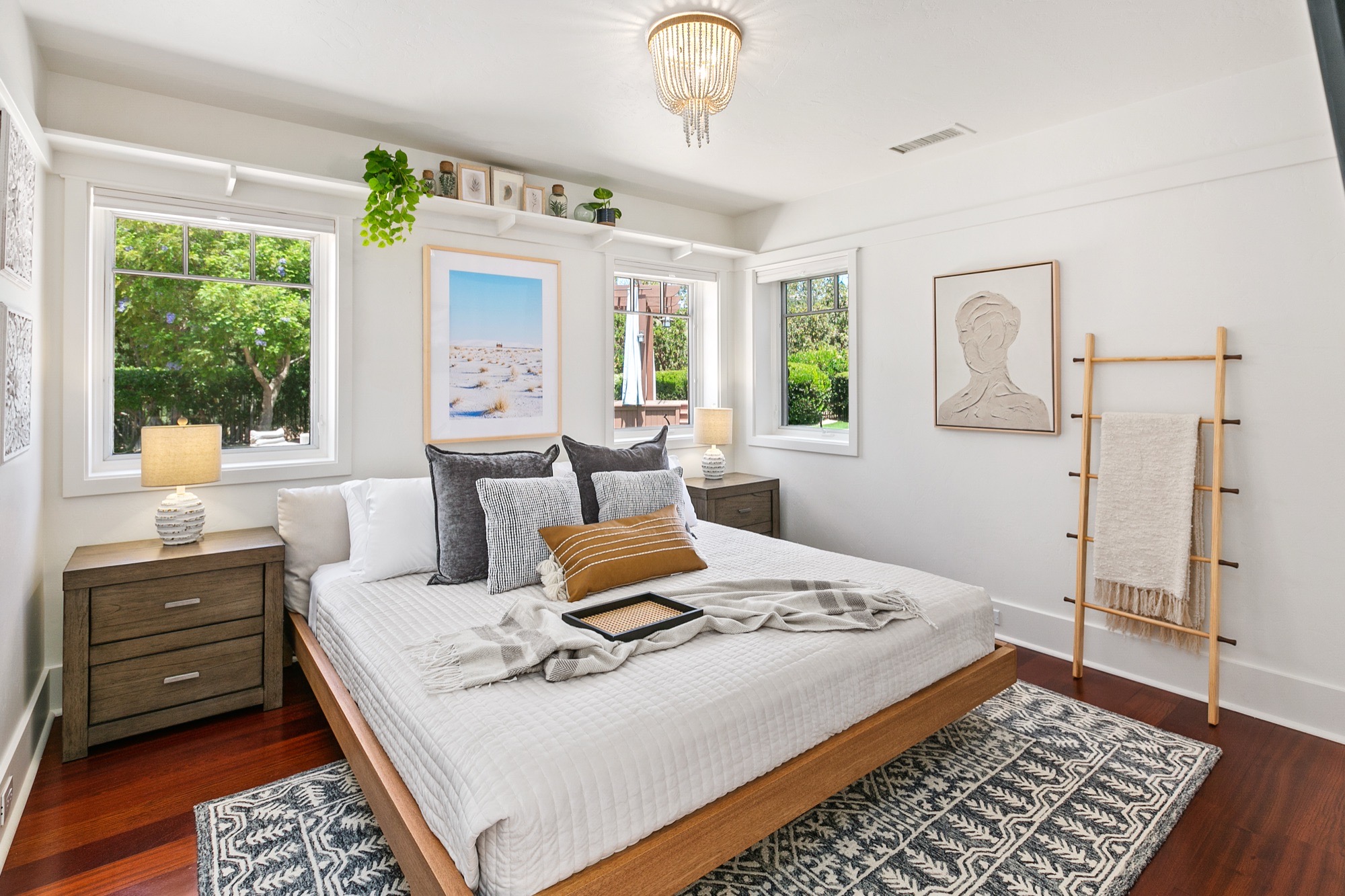 Bright white bedroom with a low wood platform bed, beaded chandelier, plant-topped picture ledge, wooden blanket ladder and black-and-white patterned rug.