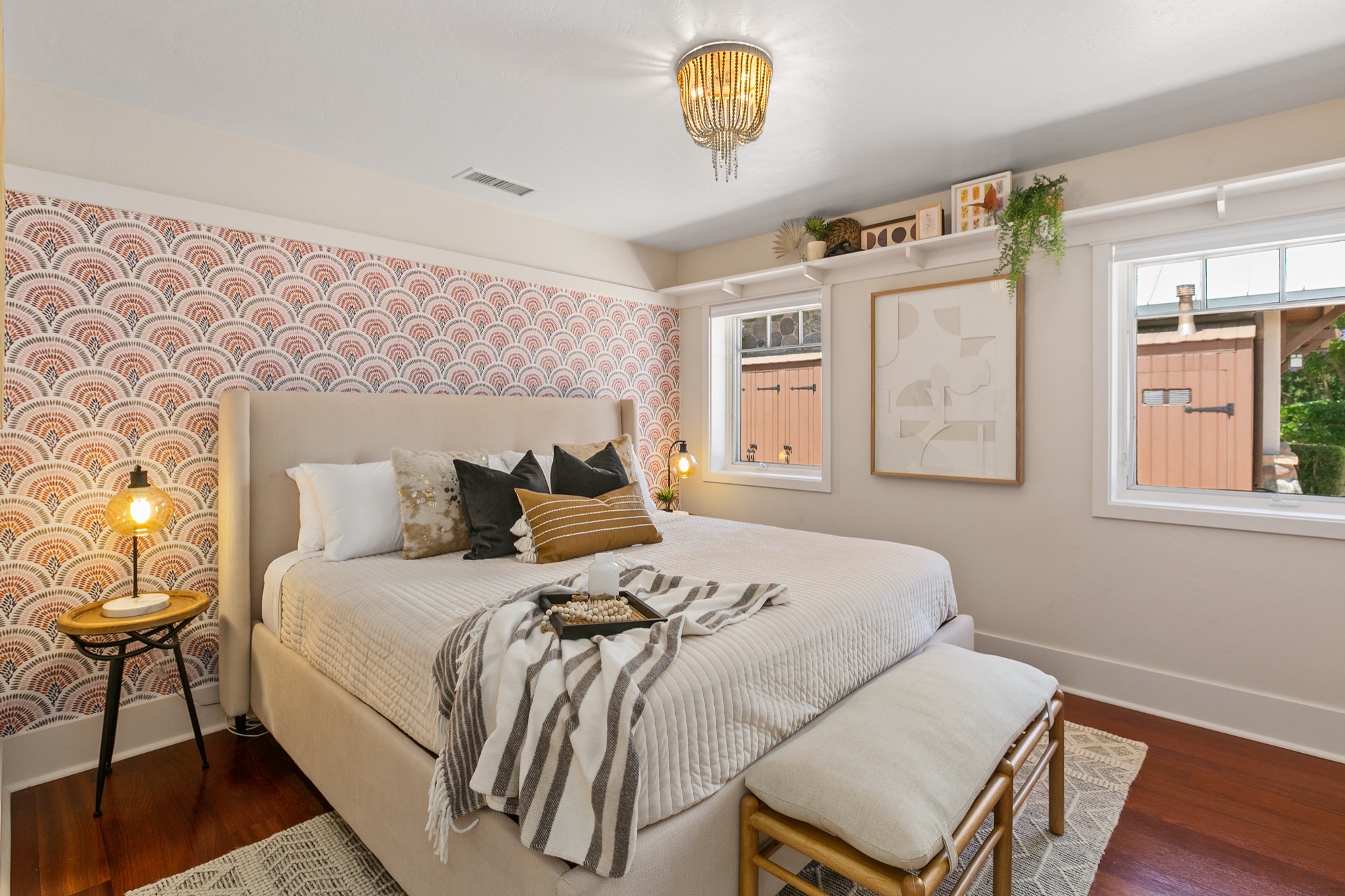 Bedroom with coral scalloped feature wallpaper, cream upholstered bed, beaded ceiling light, amber glass table lamp and cushioned bamboo-frame bench.