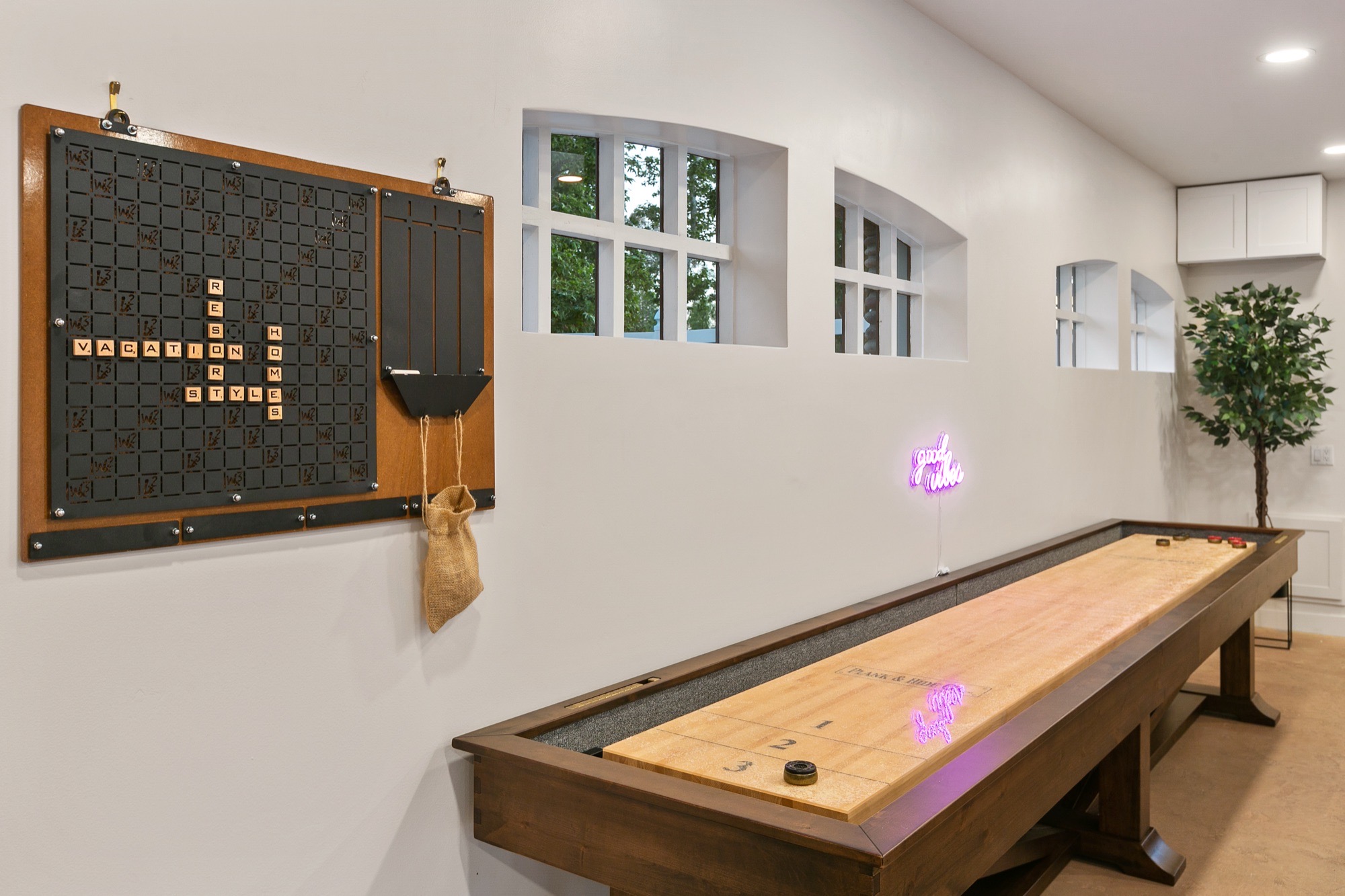 Game room detail with a long wooden shuffleboard table, wall-mounted letter-tile board, purple neon sign and arched transom windows along a white wall.