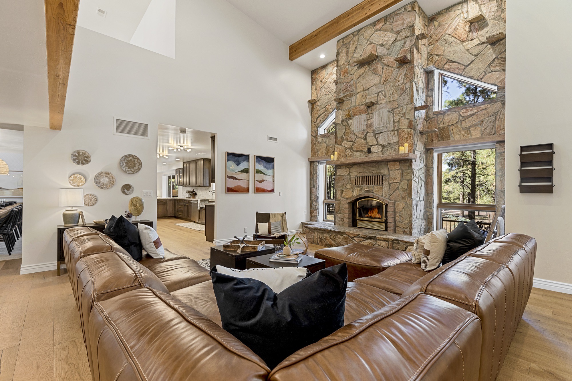 Double-height living room with a stacked-stone fireplace, timber beams, caramel leather sectional, black pillows and tall windows framing pine trees.