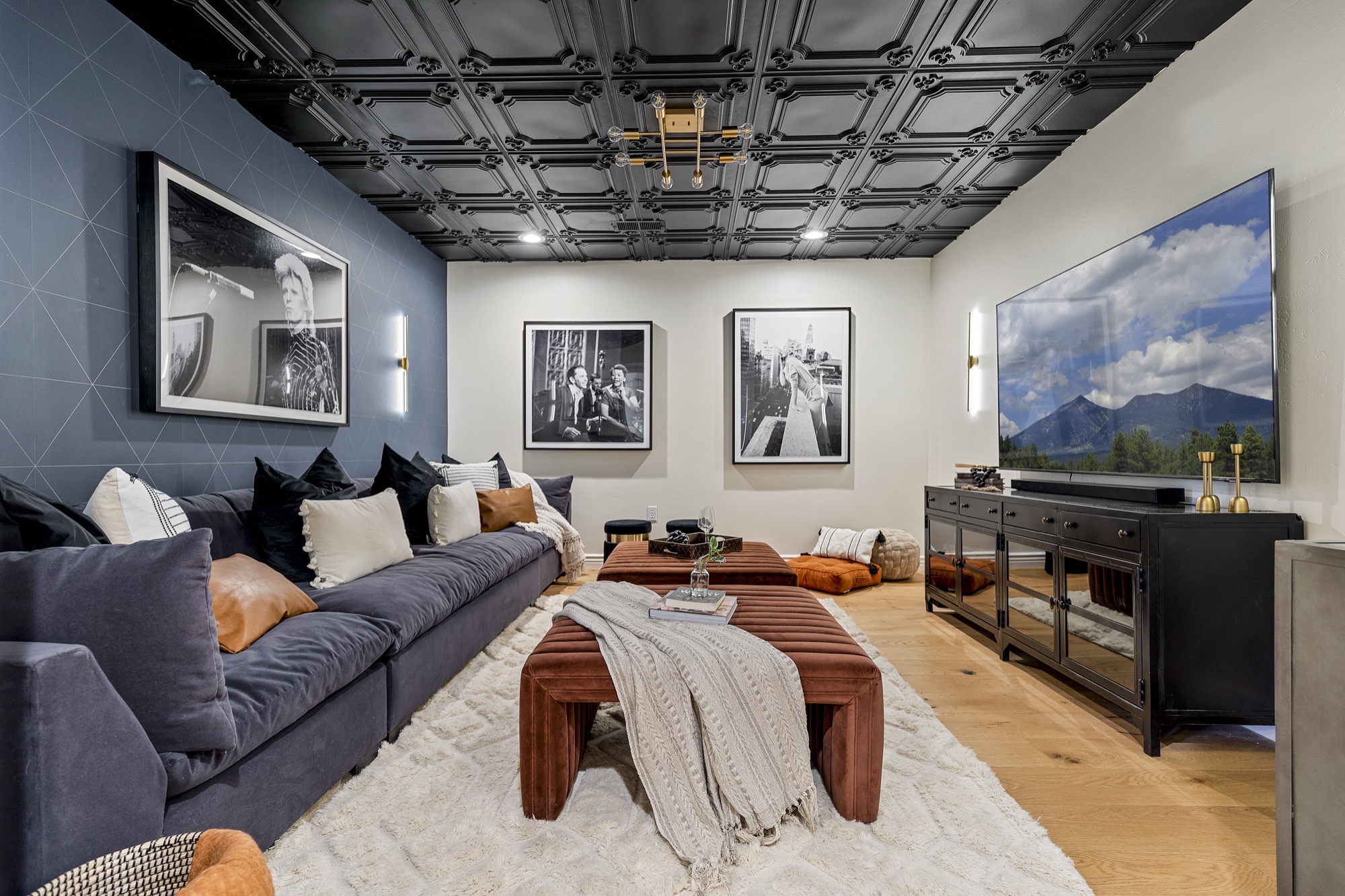 Media room with a black coffered ceiling, slate-blue geometric wallpaper, charcoal sectional, rust velvet ottomans, shag rug and black-and-white photography.