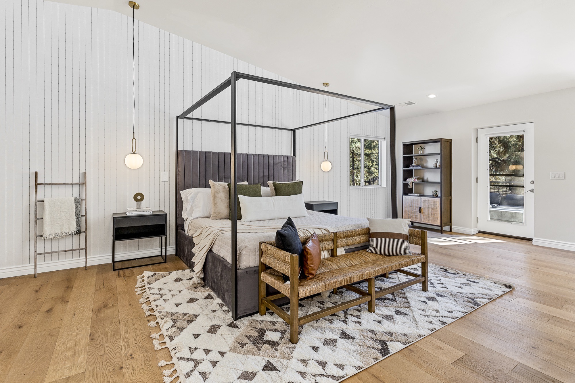Bedroom with a black metal canopy bed, charcoal channel-tufted velvet headboard, globe pendants, woven rush bench, white paneled wall and oak floors.