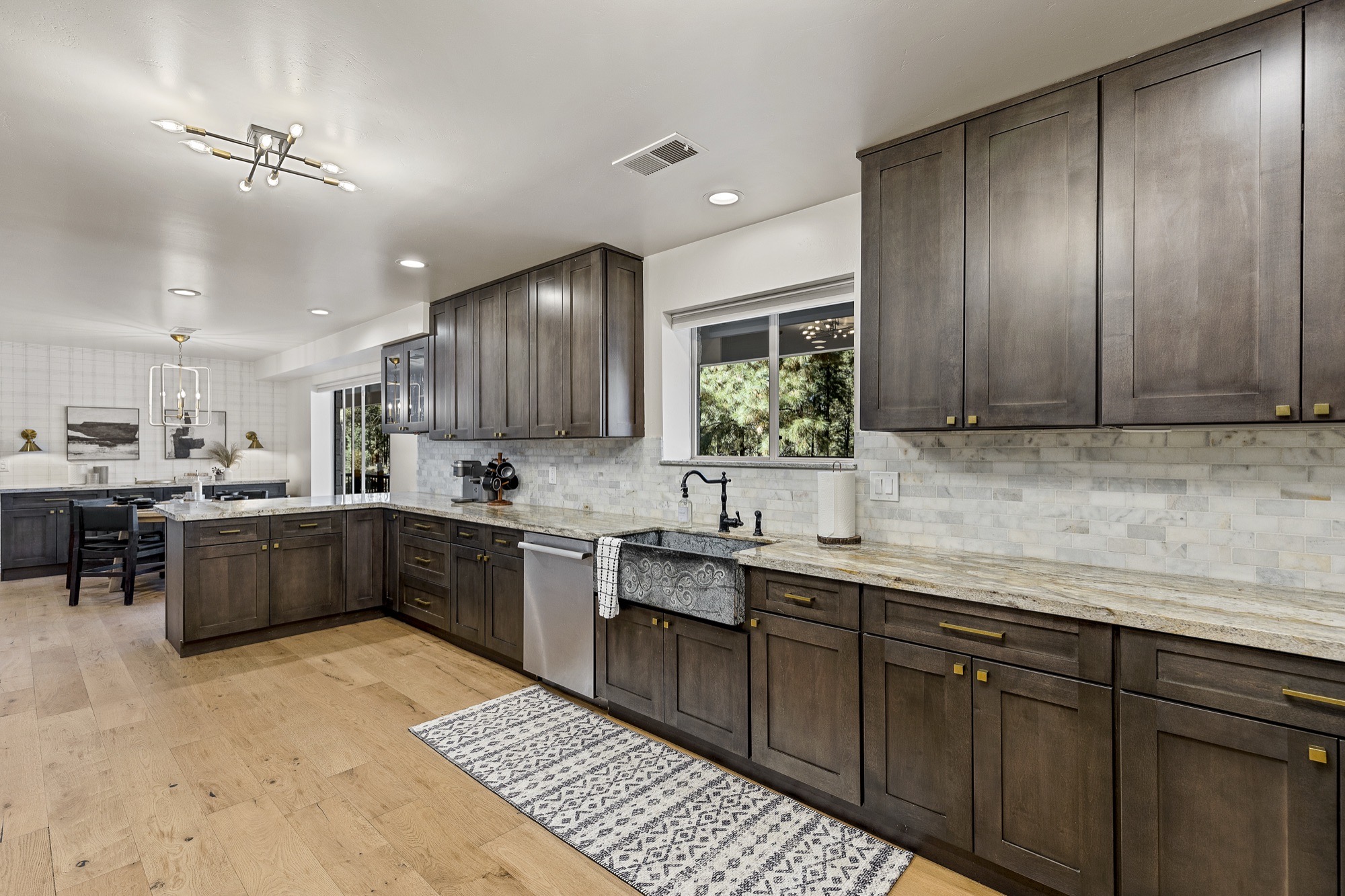Kitchen with smoky-brown shaker cabinets, brass pulls, marble subway backsplash, pale stone counters, a patterned stone farmhouse sink and oak floors.