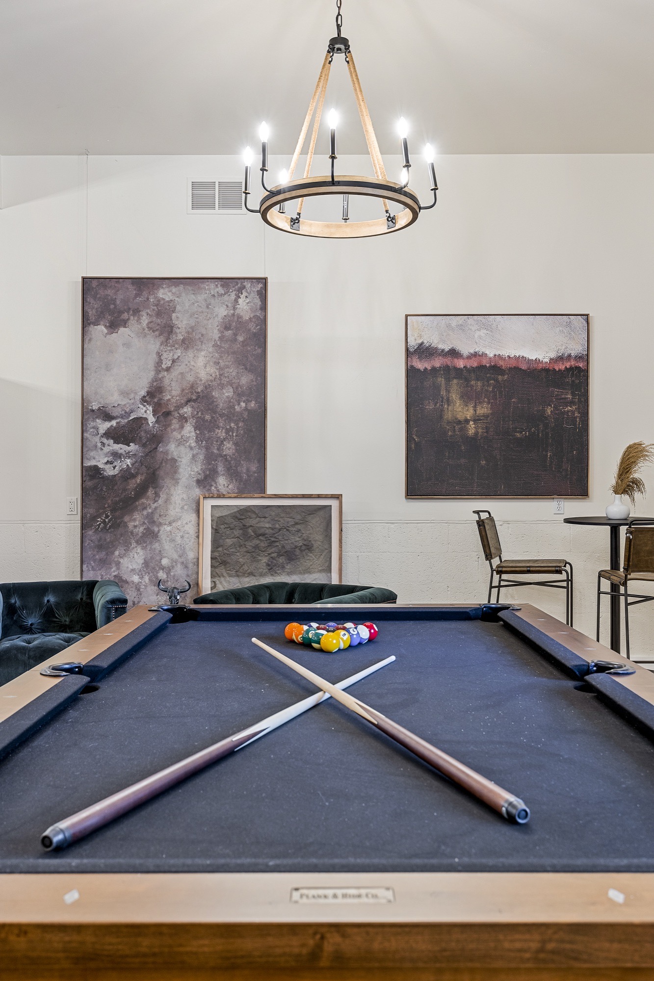 Game room with a navy-felt pool table, crossed cues, a wood-and-iron candle chandelier, large abstract canvases and green velvet chesterfield chairs.