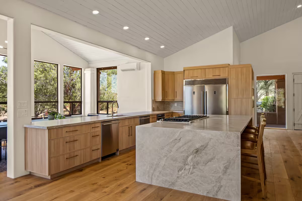 Kitchen with pale wood cabinetry, a grey veined stone waterfall island with gas cooktop, stainless refrigerator, wood counter stools and vaulted white plank ceiling.