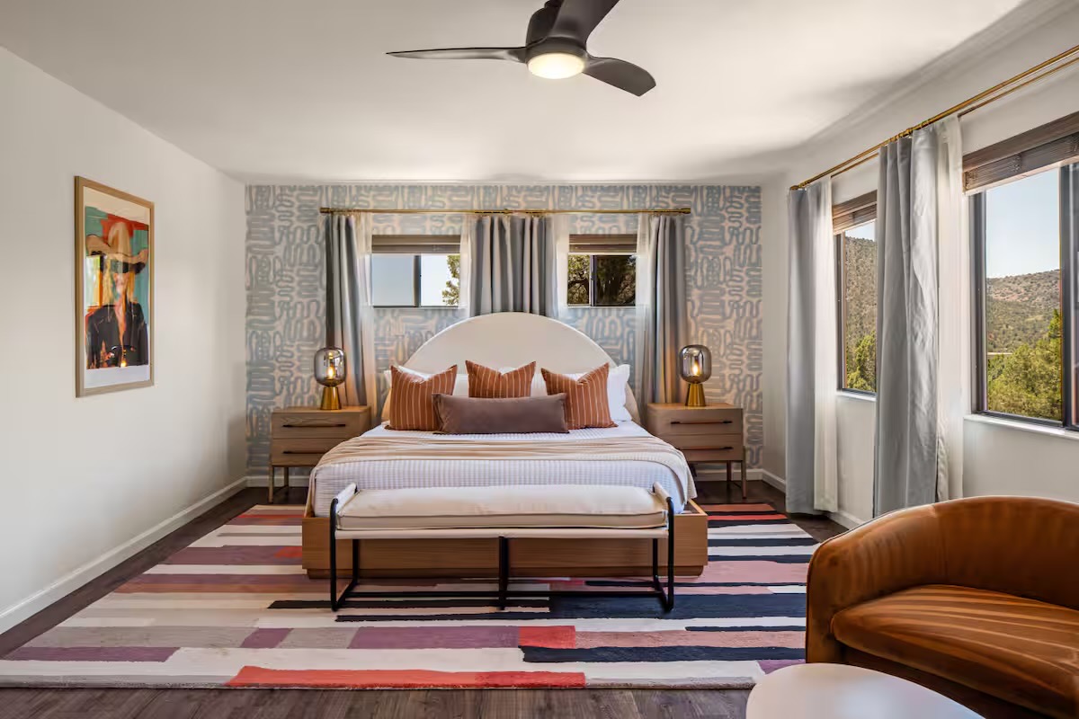 Bedroom with an arched cream headboard, rust striped pillows, blue-grey patterned wallpaper, brass lamps, a bold striped rug and a cognac upholstered armchair.