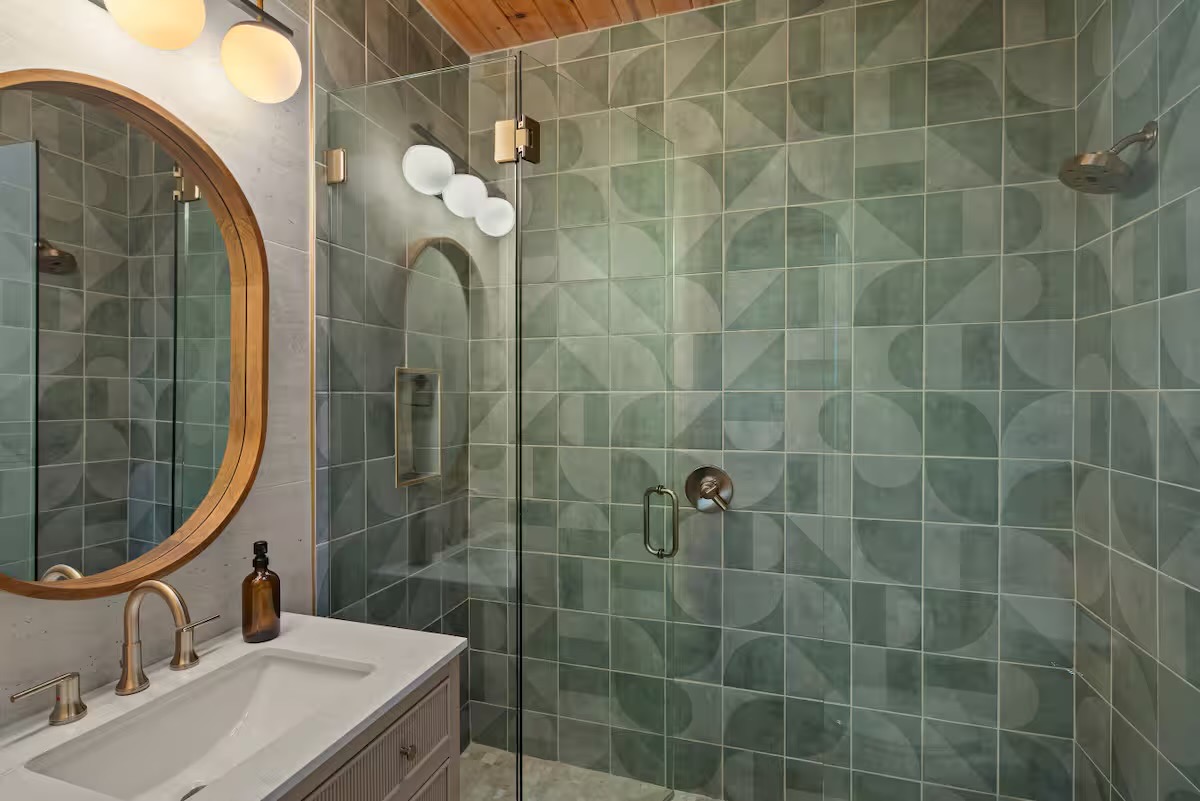 Shower room with sage green geometric tile, frameless glass enclosure, white globe lights, a rounded wood-framed mirror and brushed brass fixtures.