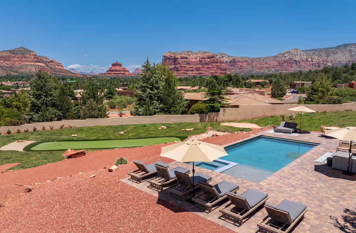 Backyard pool terrace with paver deck, grey-cushioned wood loungers, cream umbrellas, a putting green and red rock buttes beyond.