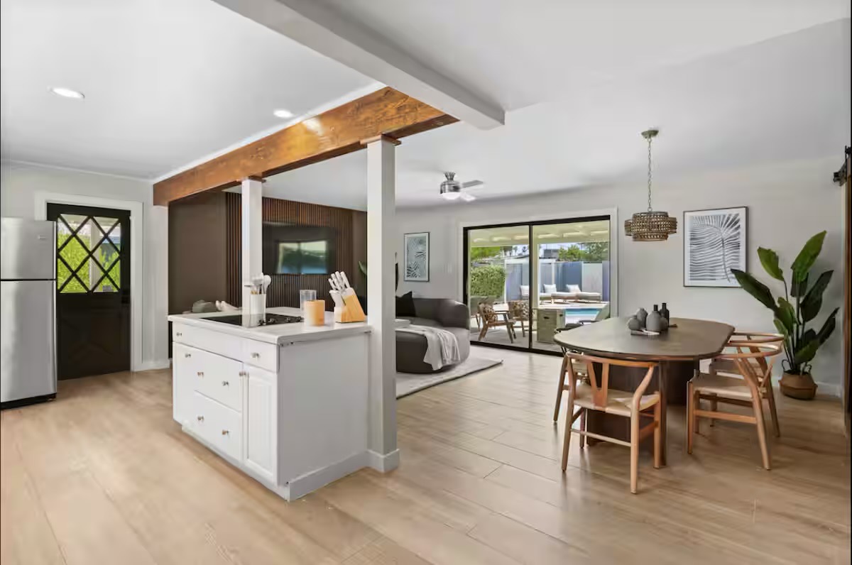 Open-plan living area with a white kitchen island under a stained timber beam, oval dining table with wishbone-style chairs, and sliding doors to the pool patio.