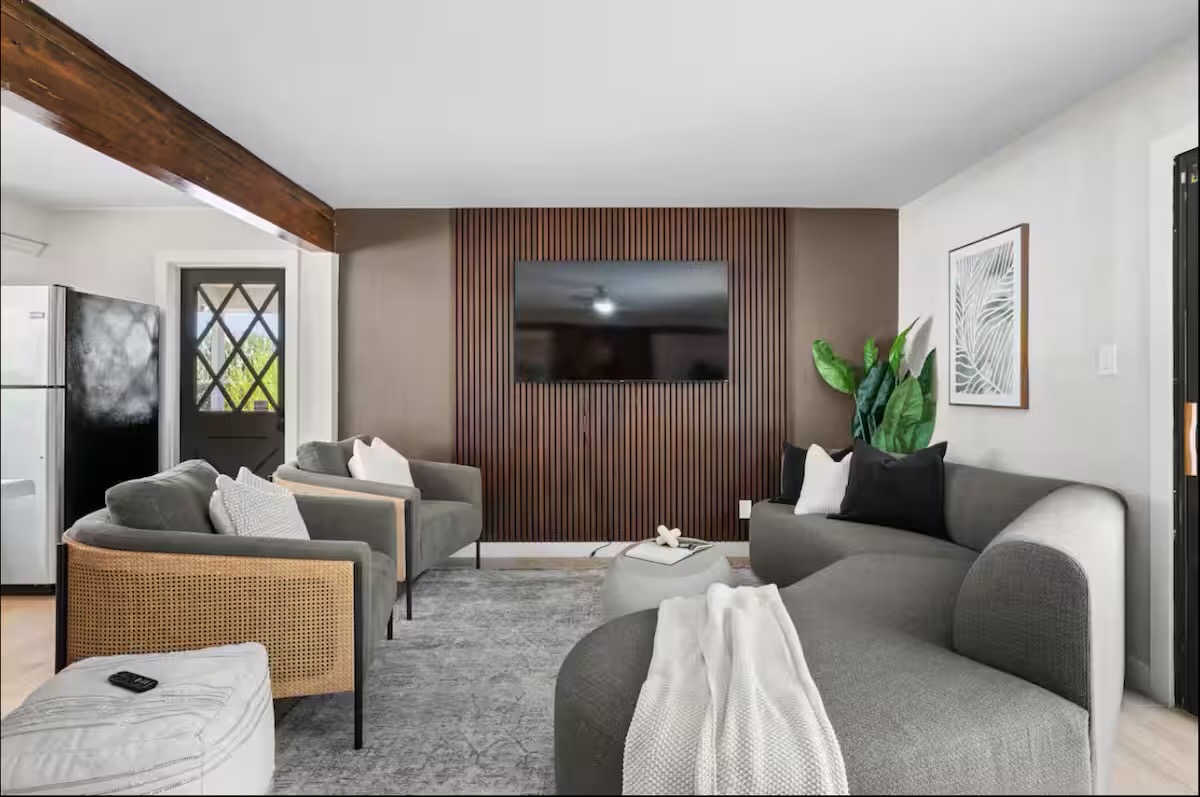 Living room with a dark timber slat feature wall, curved grey sectional, grey armchairs with woven cane sides, and a soft patterned rug.