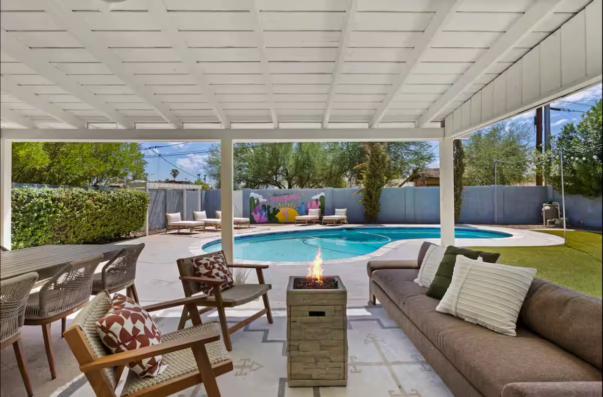 Covered patio with white-painted rafters, a long taupe sofa, woven timber lounge chairs with rust geometric pillows, and a stone-look fire table beside the pool.