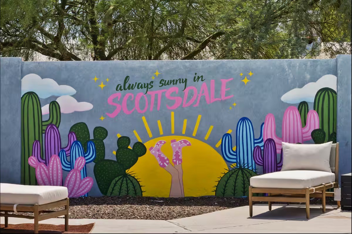 Painted mural reading 'always sunny in Scottsdale' with pink, purple and green cacti, pink cowgirl boots, a yellow sun, and cream timber loungers.