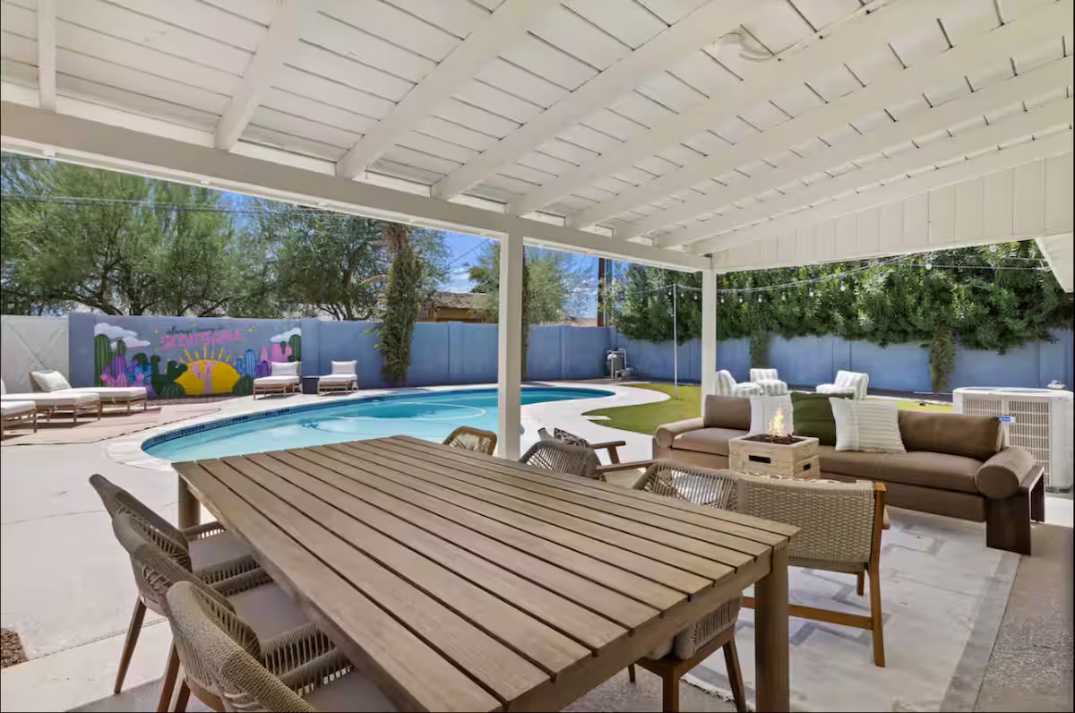 Covered patio with a long slatted timber dining table and woven chairs, a taupe lounge sofa with fire table, and the pool and mural beyond.