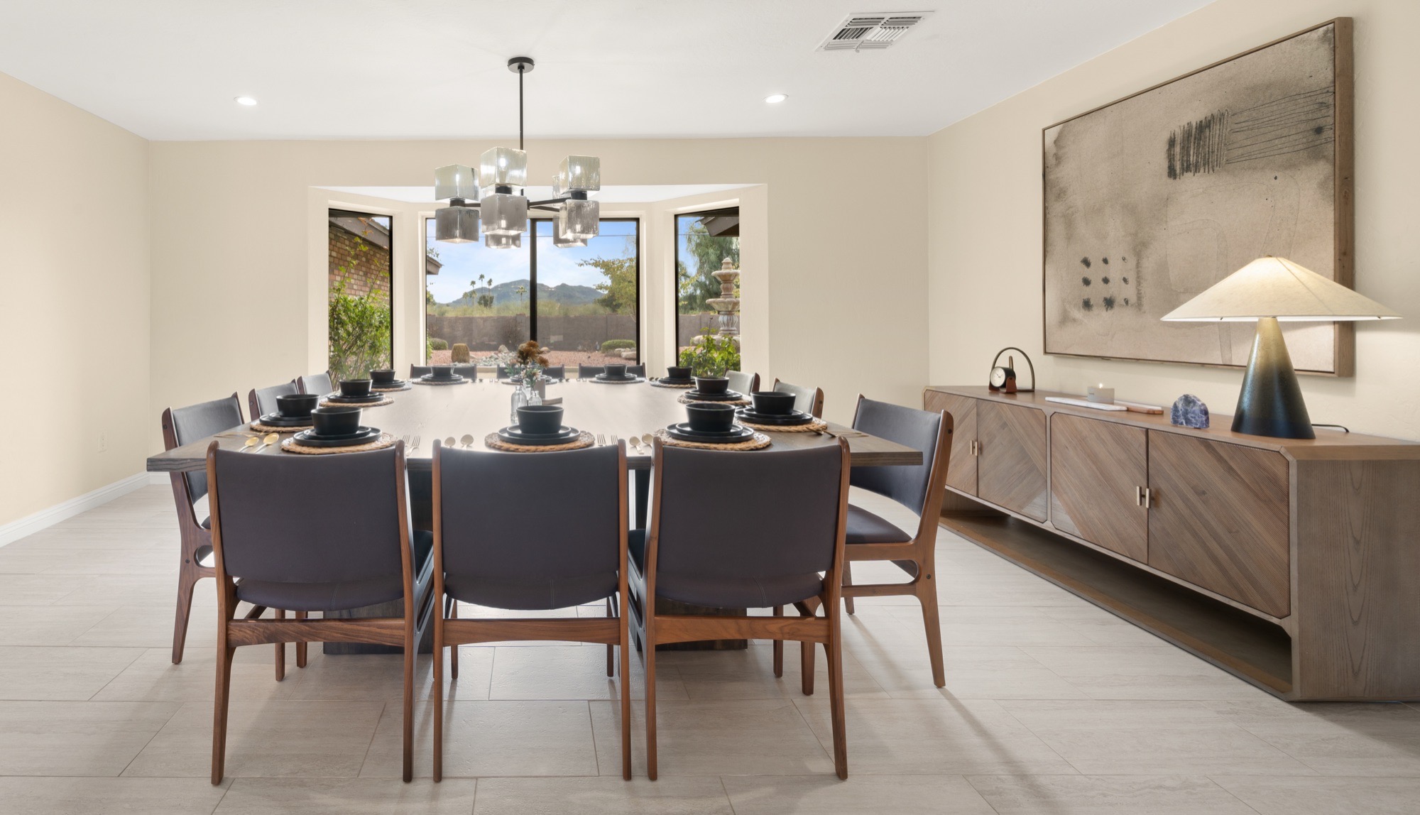 Dining room with a large square table, charcoal upholstered walnut chairs, a smoked-glass cube chandelier and a ribbed oak credenza, with a mountain view through the bay window