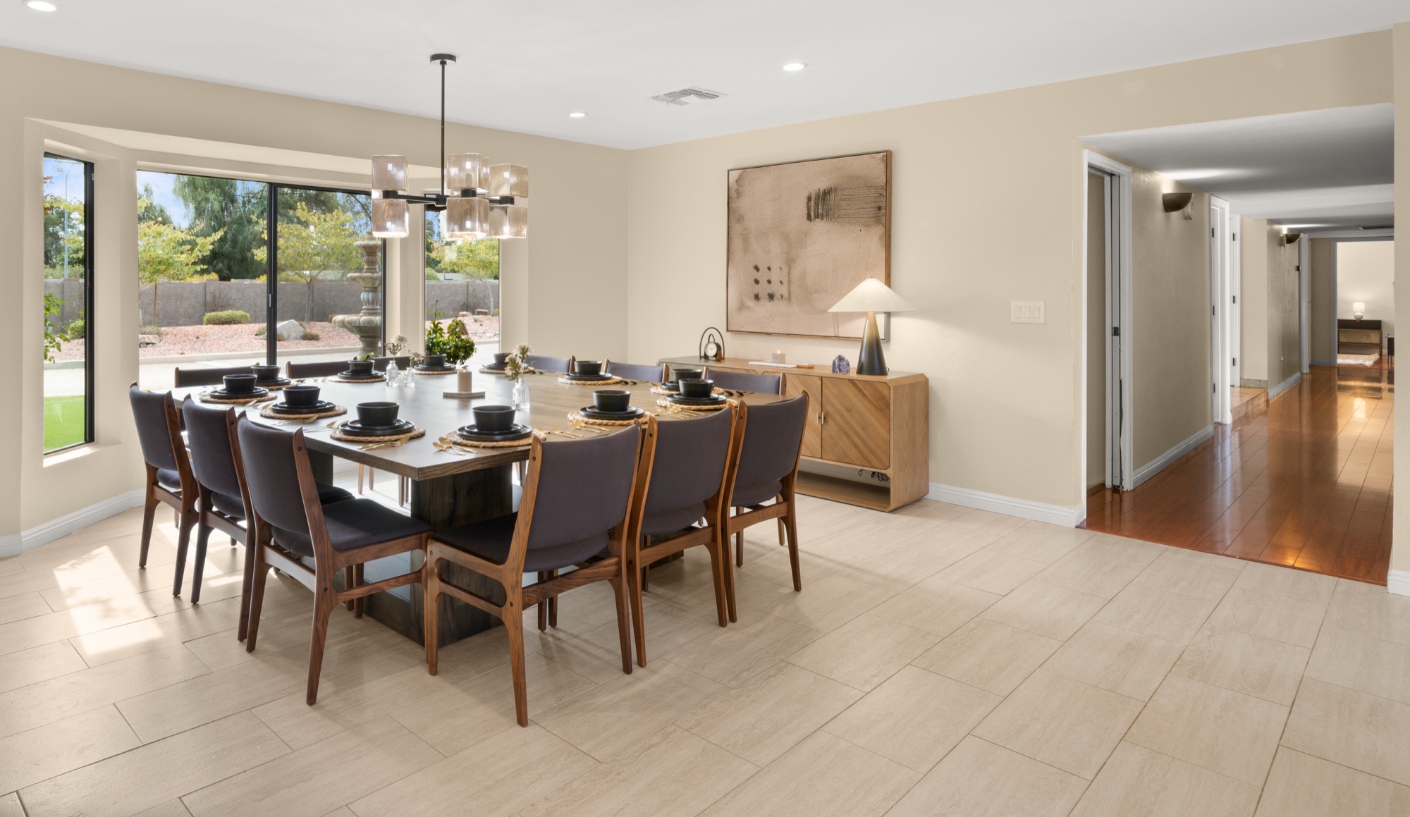 Wide view of a sunlit dining room with pale stone-look tile, a dark square table set for twelve, walnut chairs, a glass chandelier and a hallway beyond