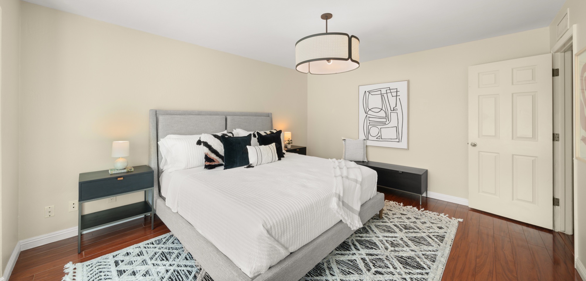 Bedroom with a grey upholstered bed, white bedding, black and white pillows, black nightstands, a linen drum pendant and a geometric black and white rug