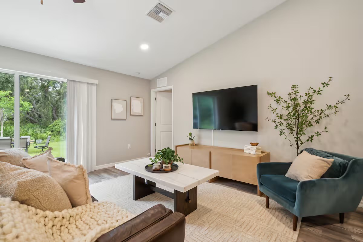 Living room under a vaulted ceiling with a teal velvet armchair, light oak media console, stone-top coffee table and sliding doors to the garden.