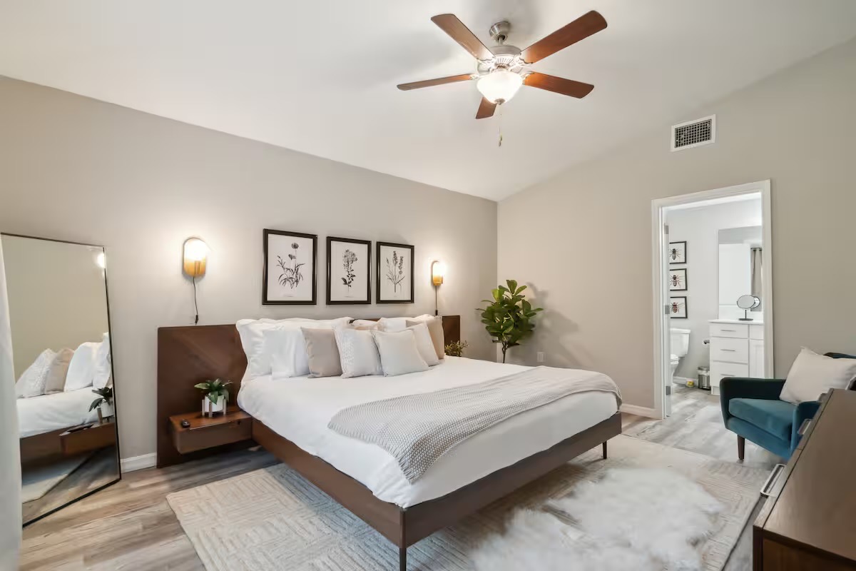 Bedroom with a walnut-toned bed and attached nightstands, white bedding, three framed botanical prints, globe wall sconces and a teal velvet armchair.