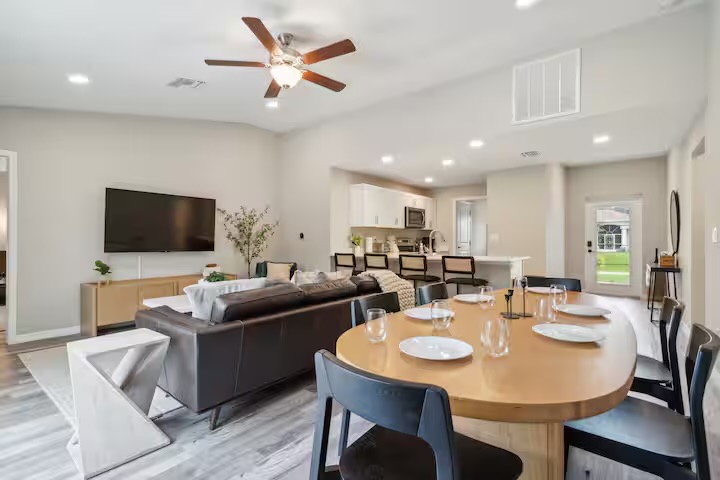 Open-plan great room with an oval oak dining table and black chairs, dark leather sofa, and white kitchen with cane-back counter stools beyond.