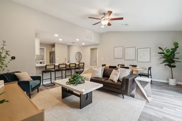 Living area with a dark leather sofa, stone-top coffee table, sculptural white side table and fiddle-leaf fig, opening to the kitchen breakfast bar.